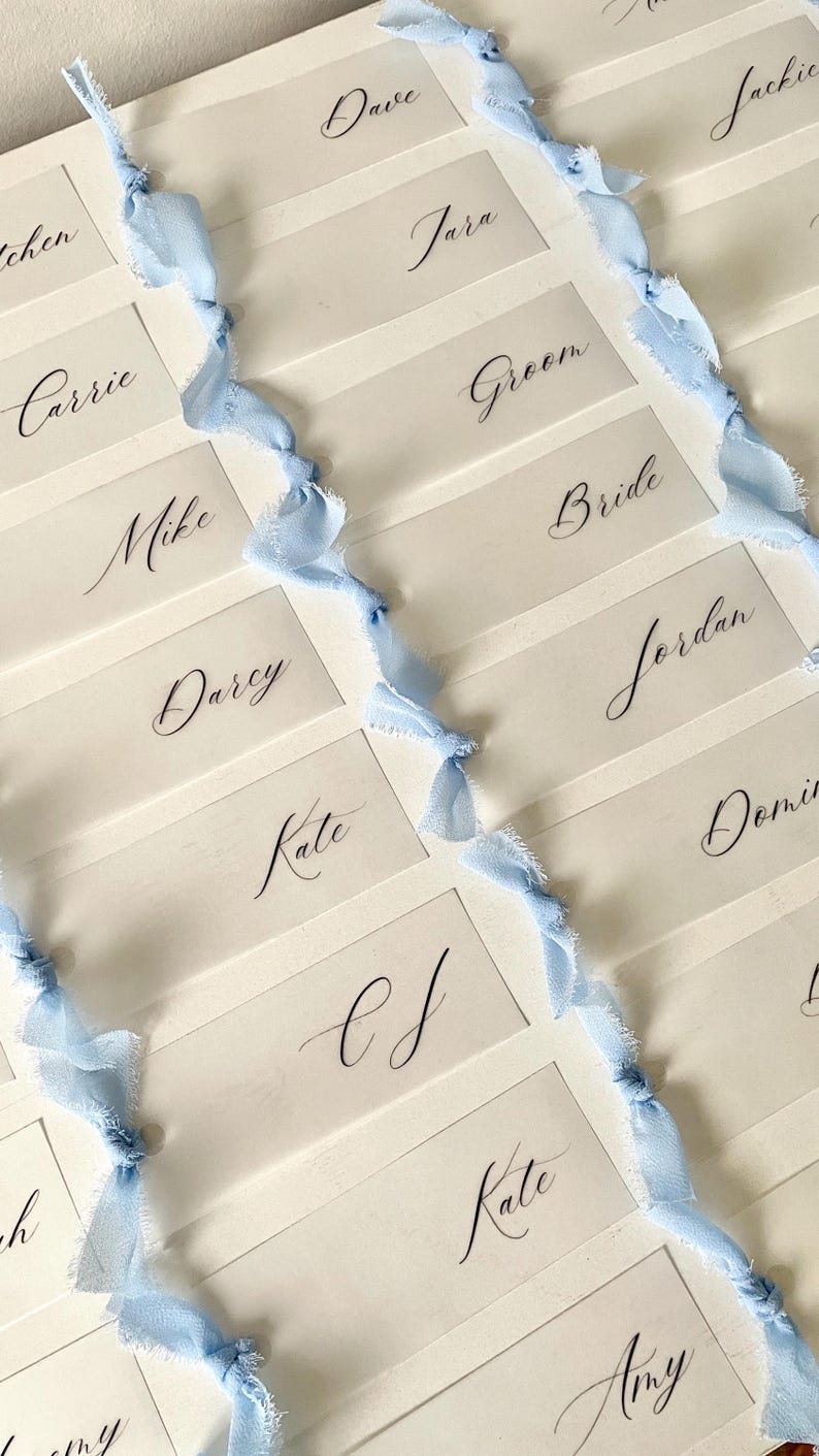 Pu&ograve; includere: Una collezione di segnaposto rettangolari bianchi con nomi in calligrafia nera come Dave, Jara, Jackie, Carrie, Groom, Mike, Bride, Darcy, Jordan, Kate e Amy. Ogni carta &egrave; decorata con una striscia verticale di nastro azzurro sfilacciato.