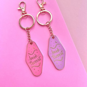 Może przedstawiać: Dwa różowe i fioletowe breloki do kluczy w kształcie klucza hotelowego z złotymi akcentami. Breloki zawierają napis "book-travert" i półksiężyc z gwiazdami.