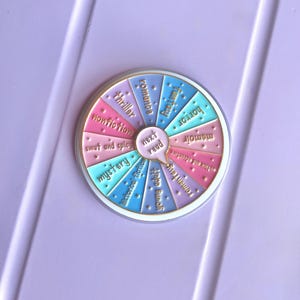 May include: A colorful enamel pin in a wheel design, featuring book genres like romance, thriller, and mystery. The pin has a central speech bubble that says "next read". The pin is set against a light purple background.