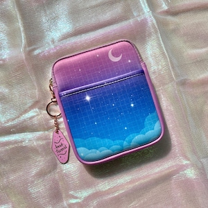 May include: A small, rectangular case with a celestial design. The case features a gradient of pink and blue, a crescent moon, and a grid pattern. A pink keychain with the text "book trove" hangs from a gold ring.