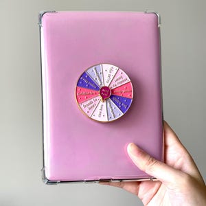 Op de afbeelding: Een roze tablethoes met een doorzichtige beschermrand, met daarin een tablet. Een kleurrijke wielspeld is bevestigd aan de tablet, met zinnen als "vrienden tot geliefden" en "date met het lot".