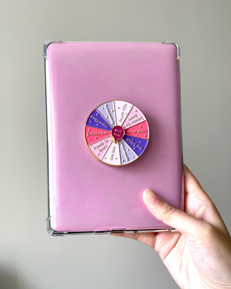 Op de afbeelding: Een roze tablethoes met een doorzichtige beschermrand, met daarin een tablet. Een kleurrijke wielspeld is bevestigd aan de tablet, met zinnen als "vrienden tot geliefden" en "date met het lot".