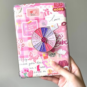 Op de afbeelding: Een roze en wit patroon tablet hoesje met diverse stickers en tekst, waaronder "Read More" en "Boss Babe". Een kleurrijke spinner is bevestigd aan de hoes, met zinnen als "Friends to Lovers" en "Take Dating".