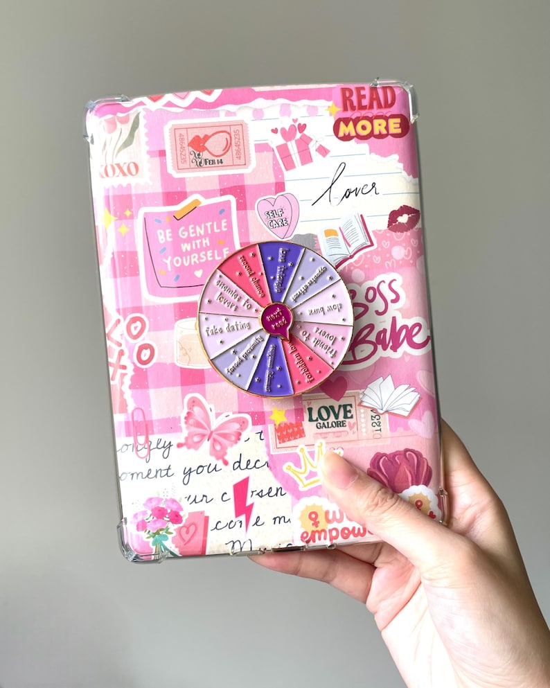Op de afbeelding: Een roze en wit patroon tablet hoesje met diverse stickers en tekst, waaronder "Read More" en "Boss Babe". Een kleurrijke spinner is bevestigd aan de hoes, met zinnen als "Friends to Lovers" en "Take Dating".