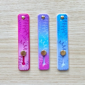 May include: Three rectangular phone grips with floral designs and gold-colored circular accents. The grips feature gradients of pink, purple, and blue. Each grip has a small, gold-colored button at the top and a circular indentation at the bottom.
