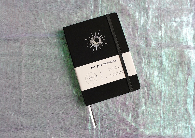 A black dot grid notebook with a white moon and sun design on the cover. The notebook has a black elastic band closure and a white label that reads 'dot grid notebook'. Dot Grid Journal, Dot Grid Journal, Travel Journal, Reading Journal, Journals and Notebooks, Junk Journal, Dot Grid Notebook, Halloween Gift