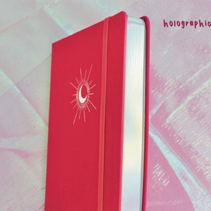 A pink hardcover journal with a white moon and sun design on the front cover. The edge of the journal is silver and has a holographic finish. Dot Grid Journal, Dot Grid Journal, Travel Journal, Reading Journal, Journals and Notebooks, Junk Journal, Dot Grid Notebook, Halloween Gift