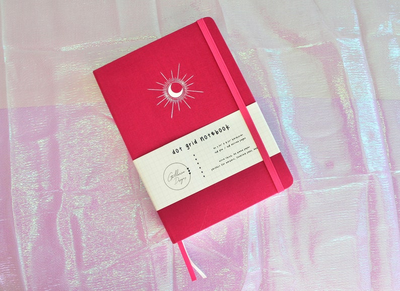A pink dot grid notebook with a white moon and sun design on the cover. The notebook has a white label that says 'dot grid notebook'. Dot Grid Journal, Dot Grid Journal, Travel Journal, Reading Journal, Journals and Notebooks, Junk Journal, Dot Grid Notebook, Halloween Gift