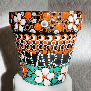 May include: Black terracotta flower pot with white and orange polka dots, white flowers, and green and blue accents.