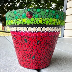May include: A tapered terracotta plant pot decorated with a dot-art pattern. The upper rim features green circles and a small ladybug, while the lower section displays red circles with dark green teardrop-shaped seeds resembling a watermelon.