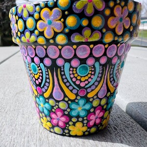 May include: A black terracotta flower pot with a colorful hand-painted design. The design features flowers, dots, and swirls in shades of pink, yellow, blue, and green.