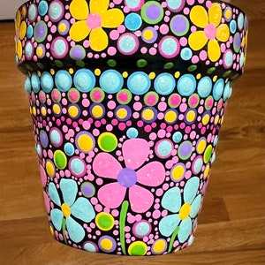 May include: A black terracotta flower pot decorated with colorful polka dots and flowers. The pot is painted with pink, yellow, blue, green, and purple dots and flowers.