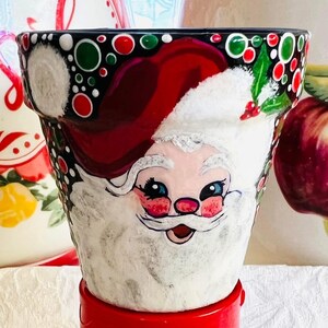 May include: A hand-painted ceramic flower pot featuring Santa Claus. The pot has a red base and a black rim decorated with red and green circles. Santa's face is painted on the front, with a white beard and a red hat.
