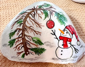 May include: A white painted rock featuring a hand-painted scene of a snowman wearing a red scarf and hat, reaching toward a red Christmas ornament hanging from a pine tree branch. The design includes green pine needles and brown branches.