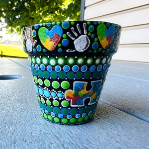 May include: A black terracotta flower pot decorated with colorful dots, a puzzle piece, handprints, and hearts. The hearts are painted with a puzzle piece design and glitter.