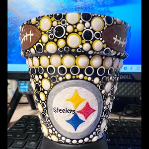 May include: A black and yellow polka dot planter with the Pittsburgh Steelers logo and the word "Steelers" painted on it. The planter has a brown football design on the top rim.