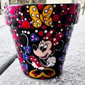May include: A hand-painted black flower pot featuring Minnie Mouse. The pot is decorated with a yellow butterfly, red, pink, and white polka dots, and blue hearts. The pot is sitting on a snowy surface.