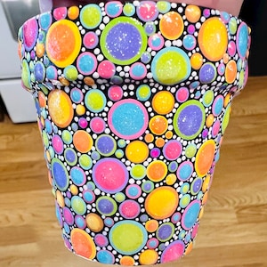 May include: A black terracotta flower pot with a colorful polka dot pattern. The dots are in various shades of pink, blue, green, yellow, and orange. Some of the dots have a glittery finish.