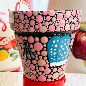 May include: A hand-painted flower pot with a black base, adorned with pink and tan polka dots. The pot features a blue oven mitt with white polka dots and a utensil design. The pot sits on a red base.