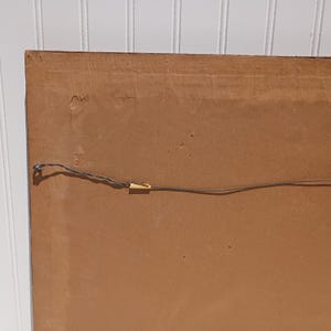 Op de afbeelding: De achterkant van een ingelijst kunstwerk, met een bruine kartonnen achterkant. Een dunne draad is over de achterkant gespannen om op te hangen. De hoek van de lijst is zichtbaar en toont de constructie.