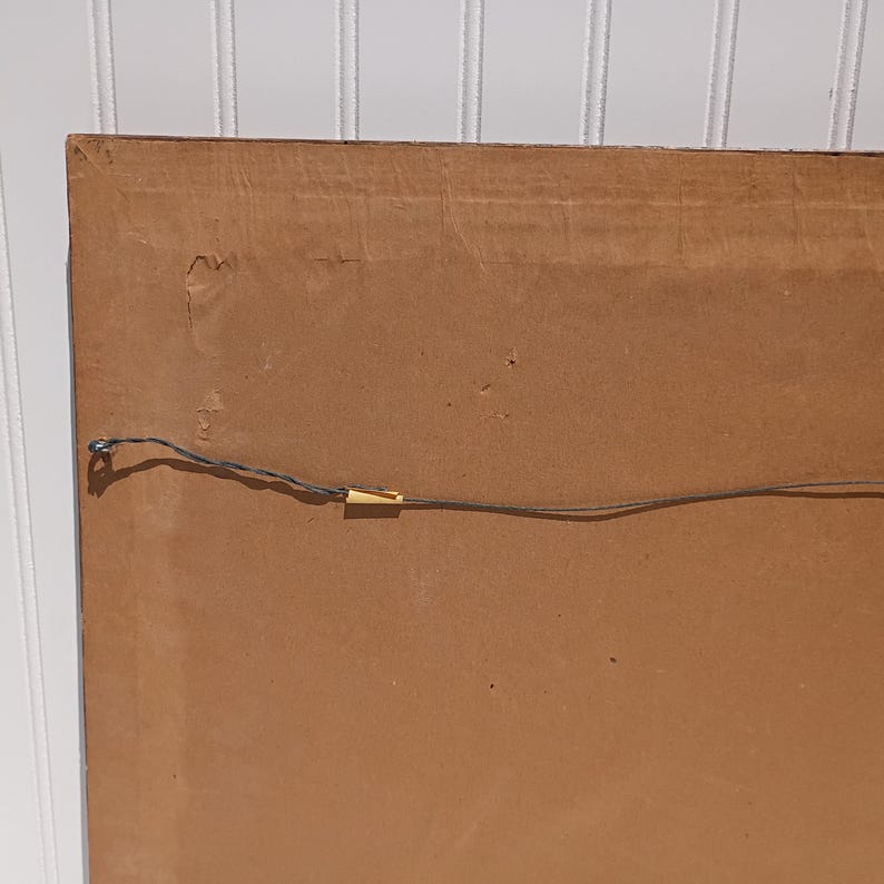 Op de afbeelding: De achterkant van een ingelijst kunstwerk, met een bruine kartonnen achterkant. Een dunne draad is over de achterkant gespannen om op te hangen. De hoek van de lijst is zichtbaar en toont de constructie.