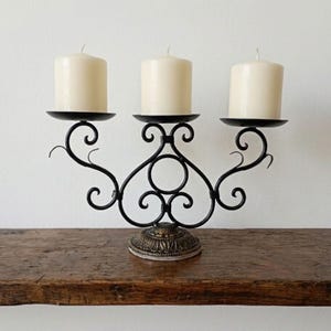 May include: A black wrought iron candelabra with three cream-colored candles. The candelabra has a decorative base and scrolled arms holding the candles. The candles are cylindrical and unlit. The candelabra is on a wooden surface.