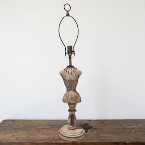 May include: A vintage-style table lamp with a mannequin torso design. The lamp features a cream-colored, distressed finish on the base and torso, with a dark metal lamp frame. The lamp is set on a wooden table against a white background.