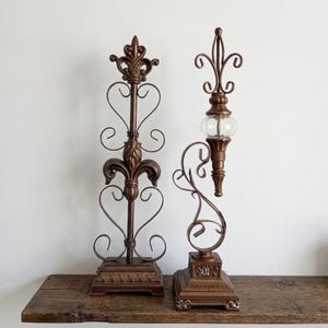May include: Two decorative bronze-colored metal candle holders. One features a fleur-de-lis design with heart-shaped accents. The other has a clear glass globe and ornate scrollwork. Both sit on square bases.