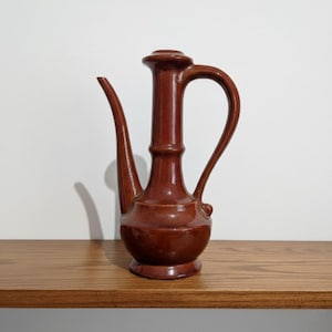 May include: A brown ceramic pitcher with a long spout and a curved handle. The pitcher is on a wooden shelf against a white wall. The pitcher has a bulbous base and a cylindrical neck.