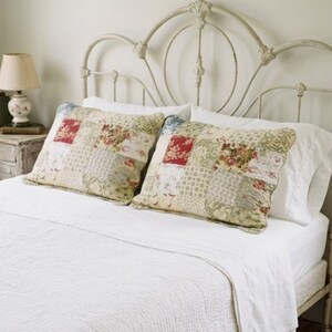May include: Two rectangular pillows featuring a patchwork design with various floral and botanical patterns in shades of red, green, cream, and blue. The pillows are placed on a white bed with a white quilt, set against a cream metal headboard.