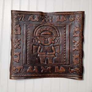 May include: A brown leather wall hanging with an embossed design. The central image is a stylized figure with a headdress, surrounded by a border of animal and geometric motifs. The piece has a square shape and a textured surface.