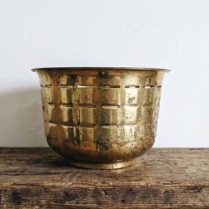 May include: A round brass planter featuring a textured grid pattern on its exterior surface. The metal has a weathered, aged patina with visible dark spots and a polished rim. The vessel has a wide opening that tapers down to a circular base.
