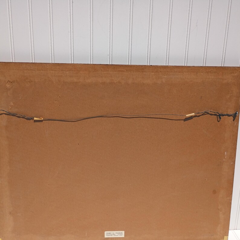 Op de afbeelding: Achterkant van een ingelijst kunstwerk met een bruine papieren achterkant. Een draad is gespannen om op te hangen. De lijst heeft een donkere houtkleur. Het kunstwerk is bevestigd met kleine metalen clips.
