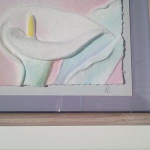 May include: A framed watercolor painting of a calla lily. The flower is white with a yellow stamen, set against a pastel background of pink, blue, and green. The artwork is signed in the lower right corner.