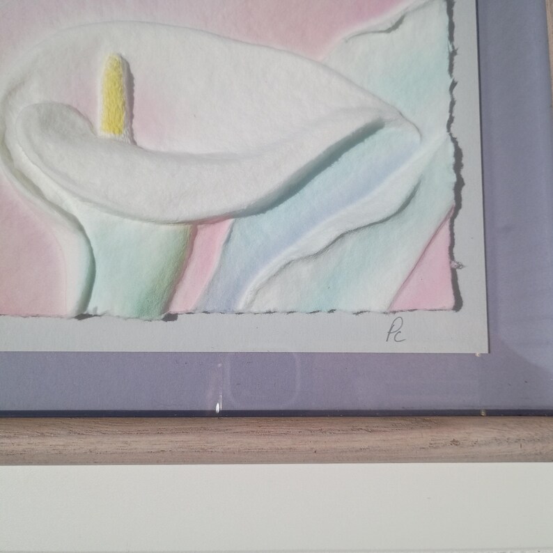 May include: A framed watercolor painting of a calla lily. The flower is white with a yellow stamen, set against a pastel background of pink, blue, and green. The artwork is signed in the lower right corner.