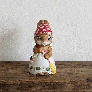 May include: A ceramic figurine of a bunny wearing a red and white checkered bonnet and a white dress. The bunny holds a broom and a small bag. The dress is decorated with green leaves and red berries. The figurine is on a wooden surface.