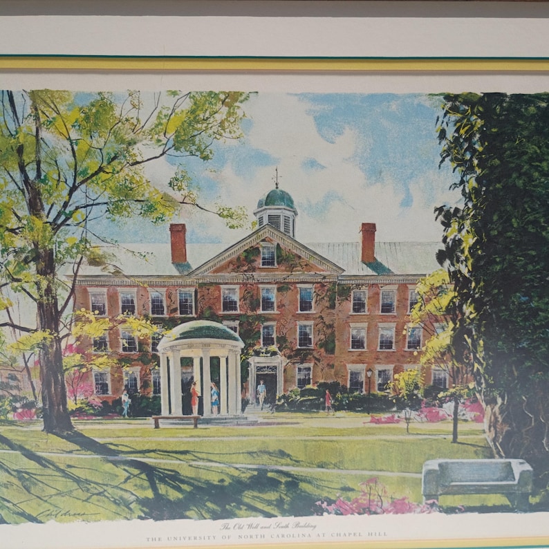 Op de afbeelding: Een ingelijste prent van de Old Well en South Building van de Universiteit van North Carolina in Chapel Hill. De prent toont een bakstenen gebouw met een groene koepel en een witte gazebo-achtige structuur. De sc&egrave;ne is omgeven door bomen en een groen gazon.