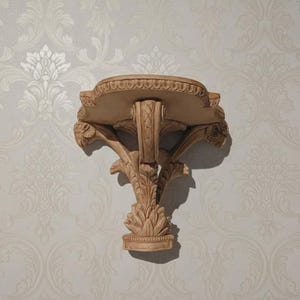 May include: A beige, ornate wall shelf with a decorative, carved design. The shelf has a rounded top with a detailed edge and is supported by a central column and stylized leaf accents. The shelf is mounted on a patterned wall.