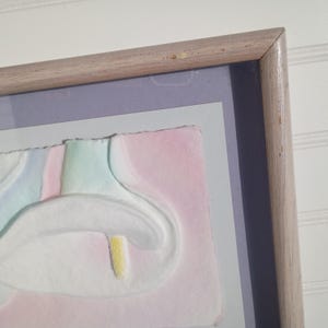 May include: A framed artwork featuring a pastel-colored floral design. The artwork has a light-colored wooden frame. The floral design includes white, pink, green, and yellow elements. The artwork is mounted on a light purple and gray background.