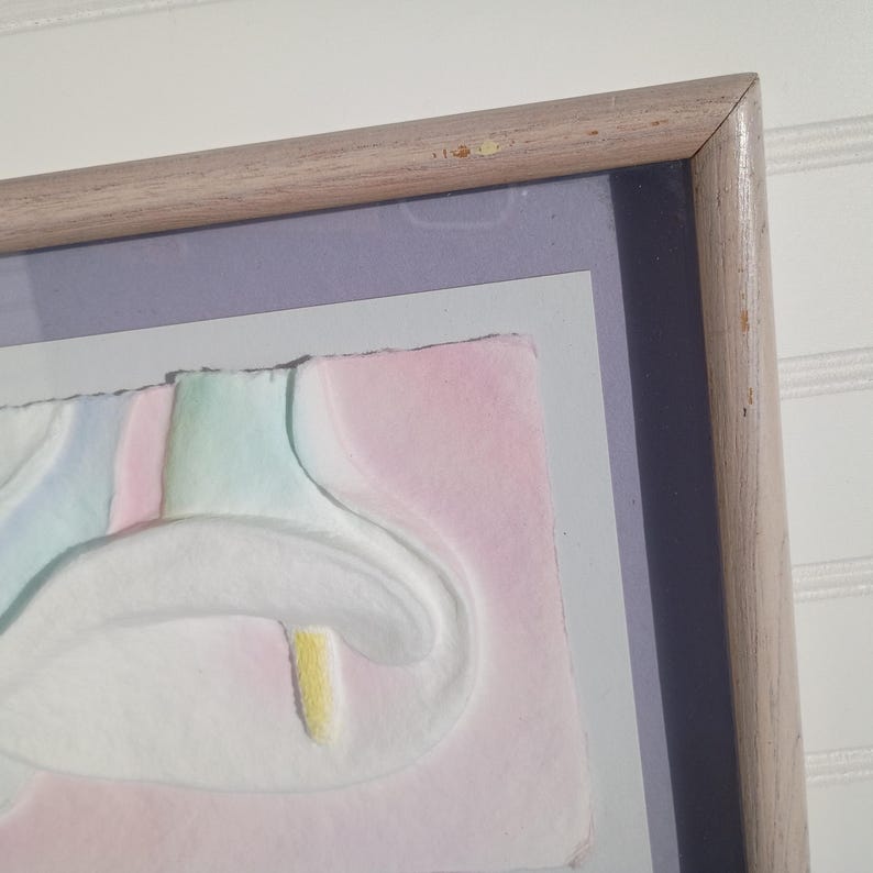 May include: A framed artwork featuring a pastel-colored floral design. The artwork has a light-colored wooden frame. The floral design includes white, pink, green, and yellow elements. The artwork is mounted on a light purple and gray background.