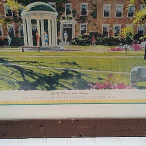 Op de afbeelding: Een ingelijste prent van The Old Well and South Building aan de Universiteit van North Carolina in Chapel Hill. De prent toont een afbeelding in aquarel van de gebouwen, mensen en het omliggende landschap. De lijst is donkerbruin.