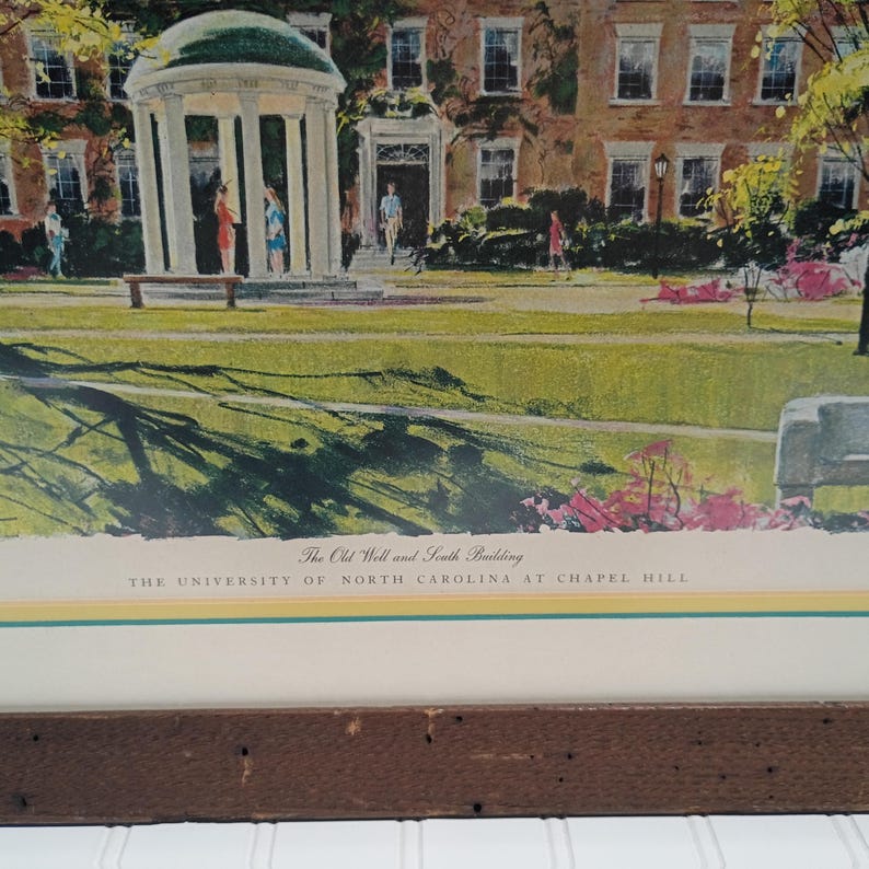 Op de afbeelding: Een ingelijste prent van The Old Well and South Building aan de Universiteit van North Carolina in Chapel Hill. De prent toont een afbeelding in aquarel van de gebouwen, mensen en het omliggende landschap. De lijst is donkerbruin.