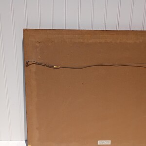 Op de afbeelding: De achterkant van een ingelijst kunstwerk, met een bruine kartonnen achterkant. Een dunne draad is over de achterkant gespannen, vastgehouden door kleine metalen bevestigingen. De rand van het frame is zichtbaar, met een houten uitstraling.