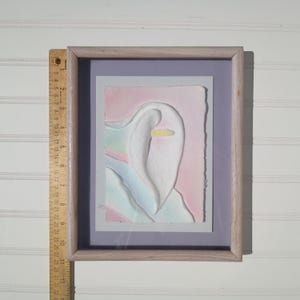 May include: A framed relief artwork featuring a white calla lily shape on a pastel pink and blue background. A wooden ruler placed alongside shows the frame height is approximately 12 inches. The artwork is set within a light-toned wooden frame.