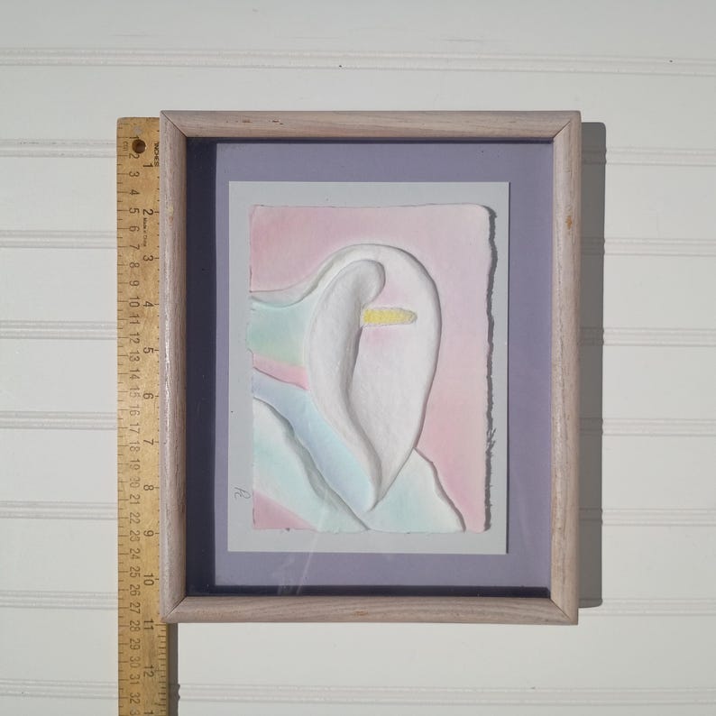 May include: A framed relief artwork featuring a white calla lily shape on a pastel pink and blue background. A wooden ruler placed alongside shows the frame height is approximately 12 inches. The artwork is set within a light-toned wooden frame.