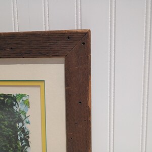 Op de afbeelding: Een ingelijst kunstwerk met een donkerbruine houten lijst. Het kunstwerk heeft een cr&egrave;mekleurige passe-partout met een gele rand. De afbeelding in de lijst toont een groen en blauw bloemenontwerp. De lijst heeft zichtbare houtnerf en spijkergaten.