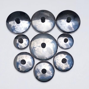 May include: A collection of nine circular, polished stainless steel pot lids arranged in a cluster. Each lid features a reflective, mirror-like surface showing sky reflections and a small, centered black knob for lifting and handling.