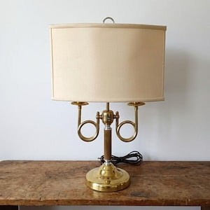 May include: A brass table lamp with a cream-colored rectangular lampshade. The lamp features a decorative base and two curved horn-like accents. The lamp is sitting on a wooden surface.