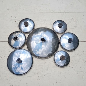 May include: A collection of seven circular, polished stainless steel pot lids of varying sizes, each featuring a small, centered black knob handle. The reflective surfaces of the lids show a clear sky with clouds, arranged in a cluster.