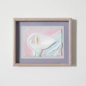 May include: A framed artwork featuring a textured, embossed paper relief of a white calla lily with a yellow spadix. The flower is set against a soft pink and light blue background with deckled edges, enclosed in a light wood frame with a lavender mat.
